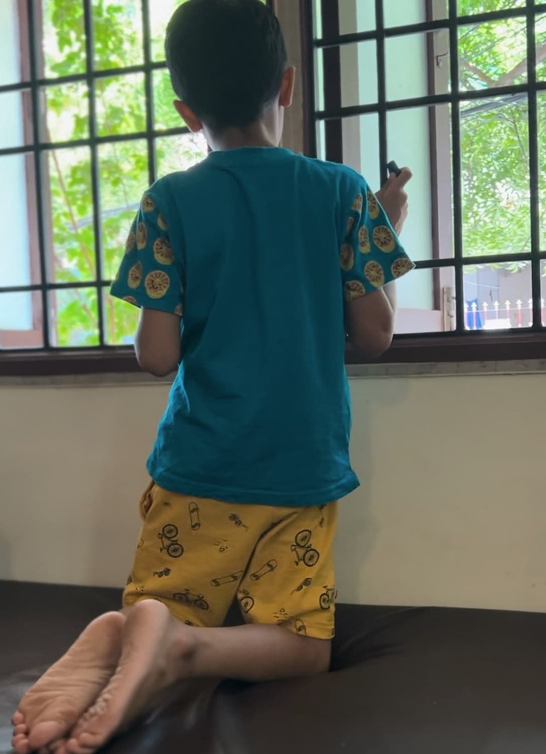 A child kneeling on a therapy bench at an open window, looking out at the trees.