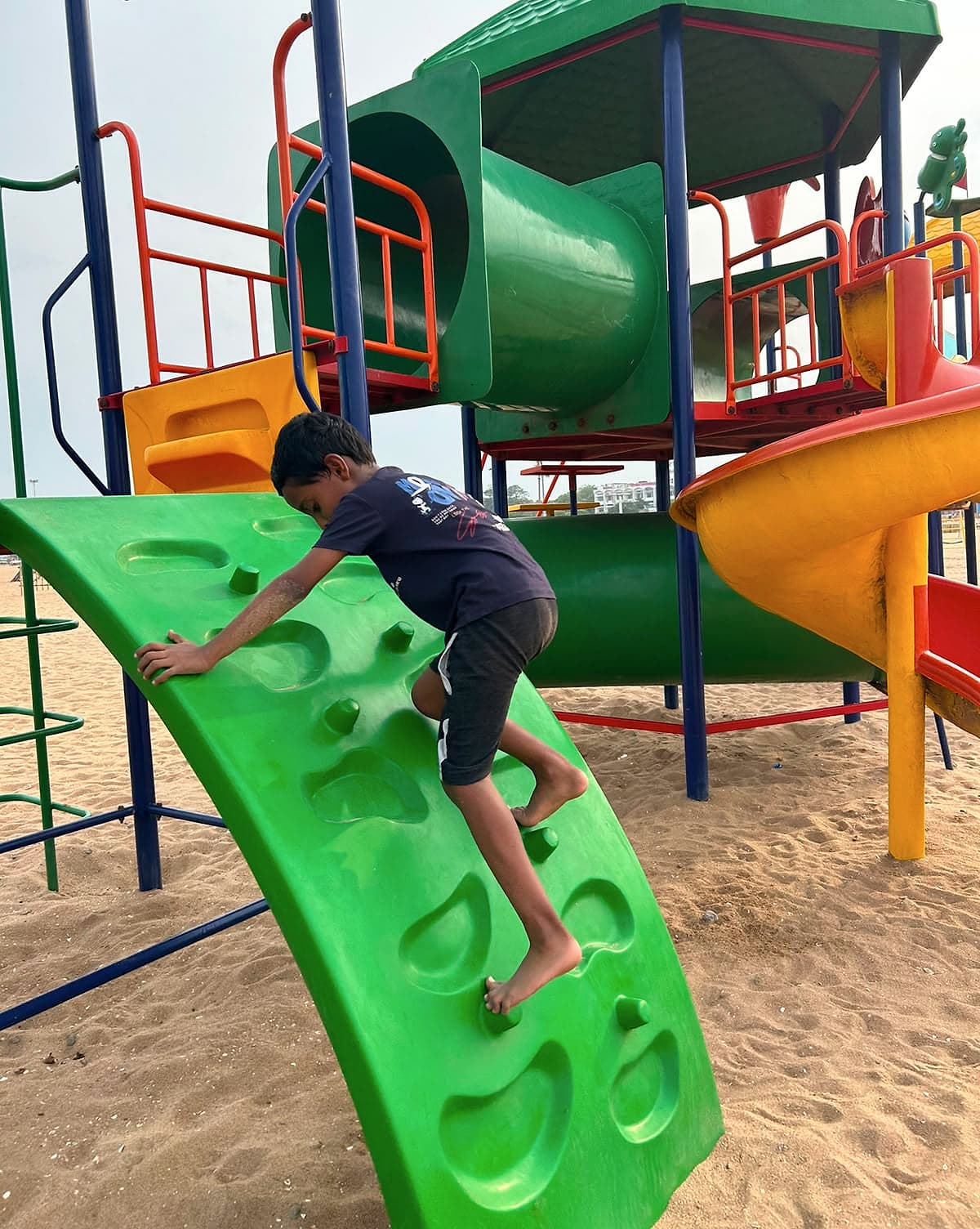 A boy climbing a green climbing wall on a playground set in the sand.