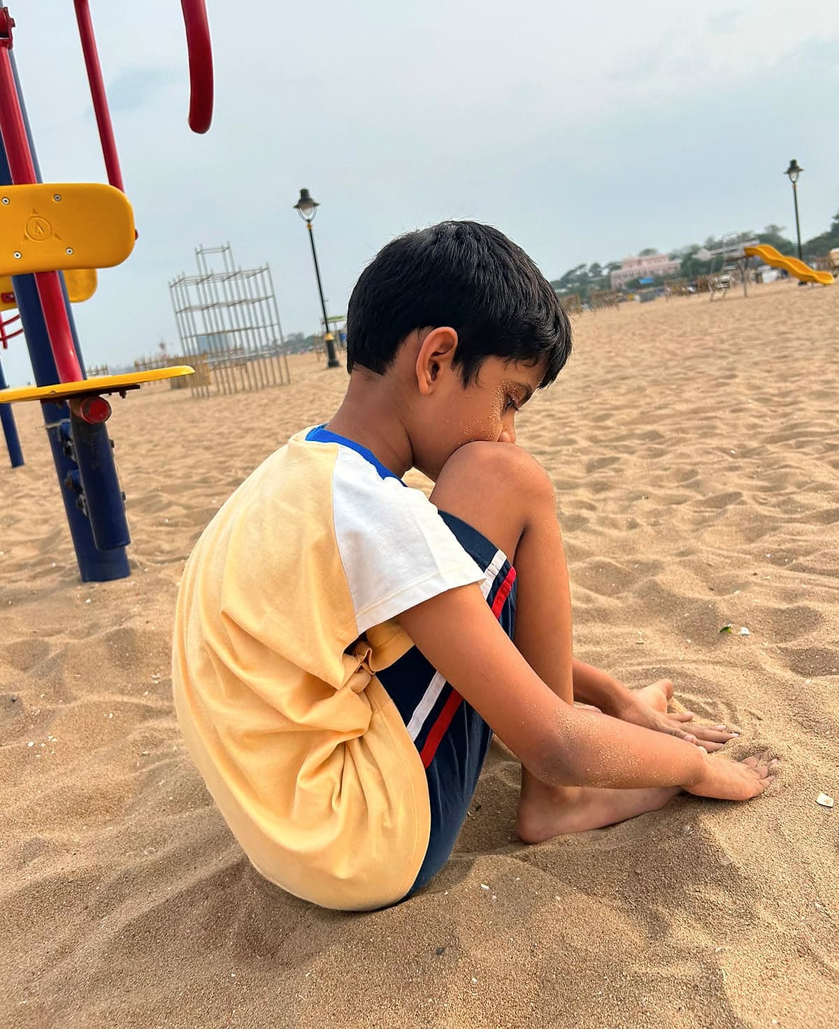 A boy sitting in the sand with his chin on his knee, his hands resting in the sand.