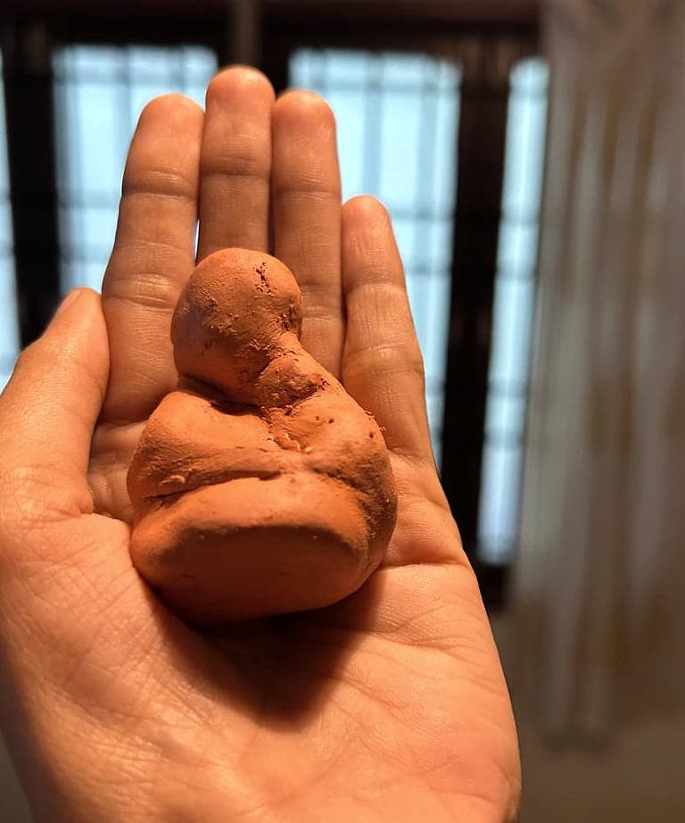 An open palm holding a small seated figure modelled in terracotta clay.