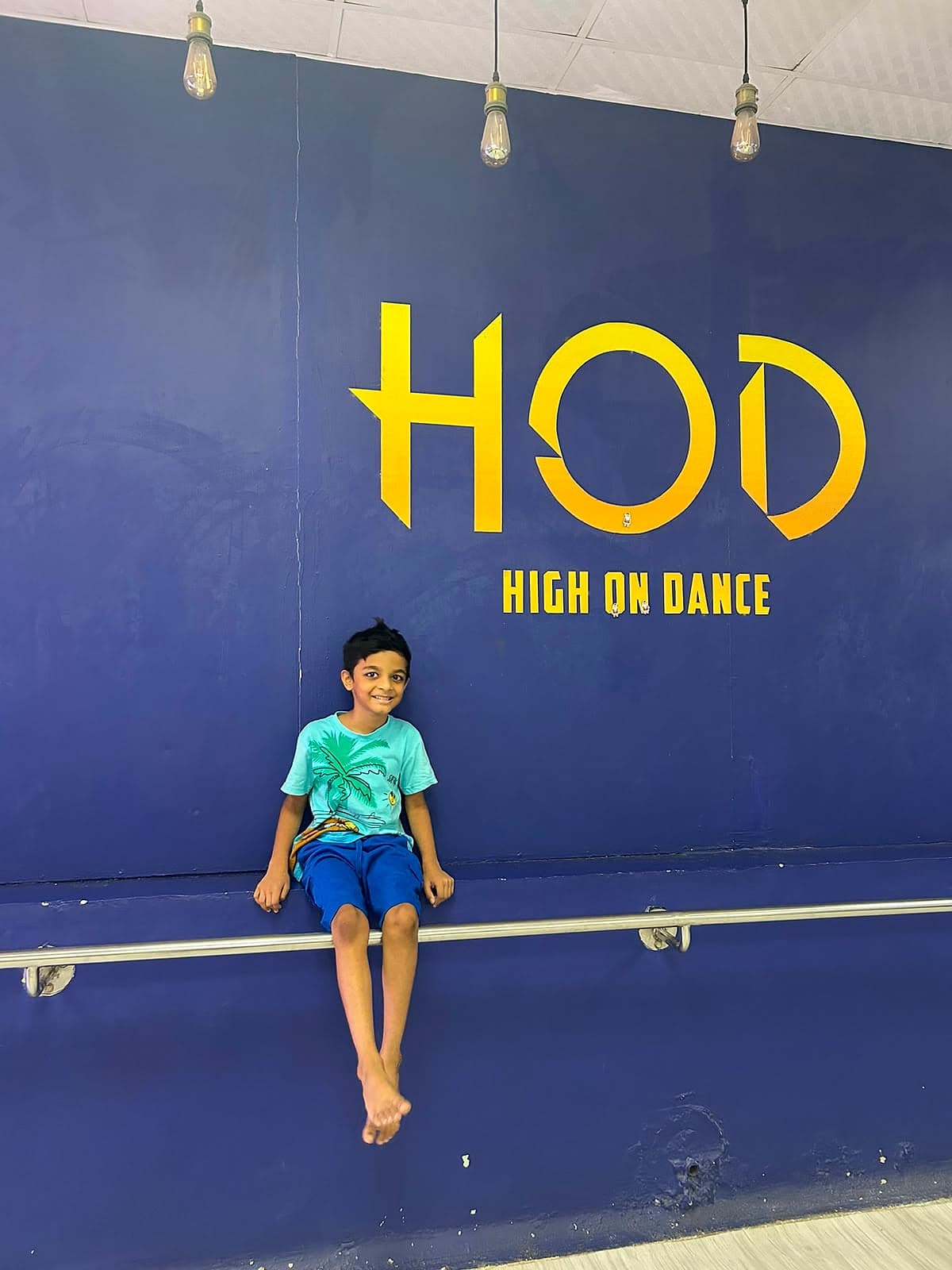 A boy sitting on a ballet barre against a dark blue studio wall, smiling.