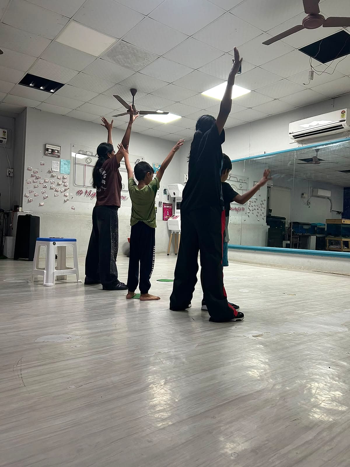 Two children and two adults in a dance studio, arms raised above their heads, seen from behind.