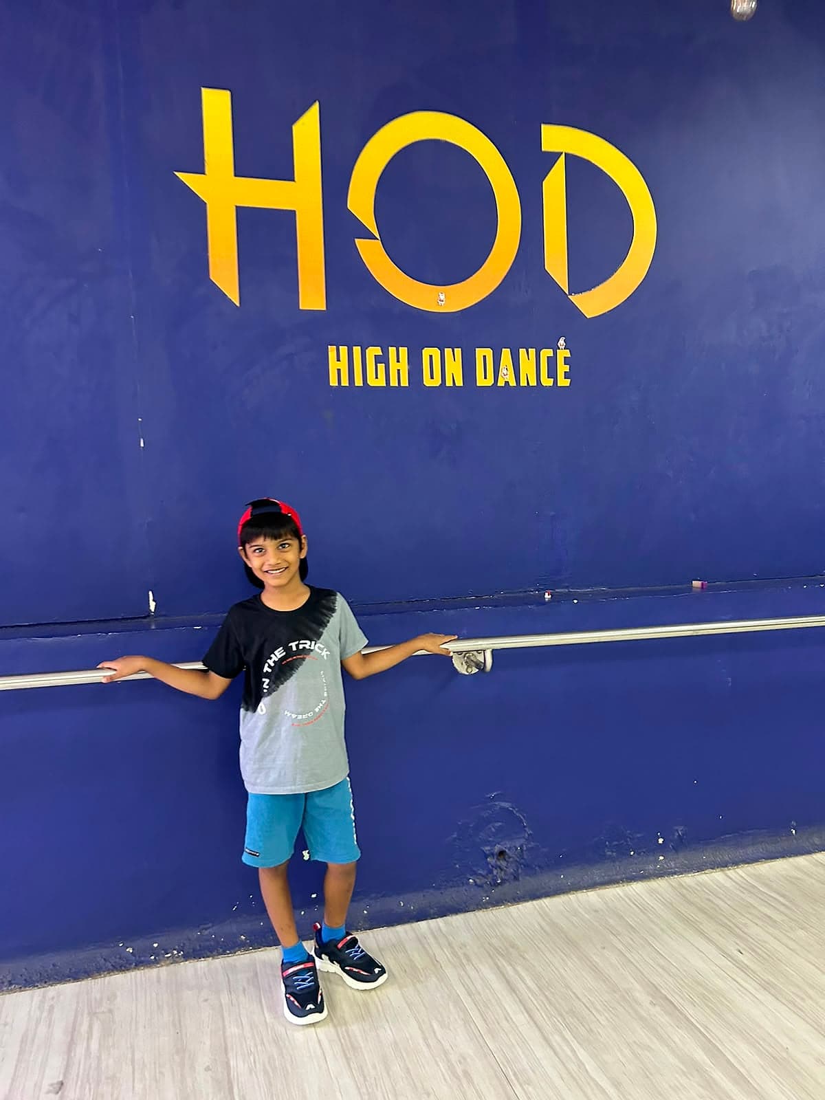 A boy in a cap standing at a ballet barre in front of a dark blue studio wall, smiling.