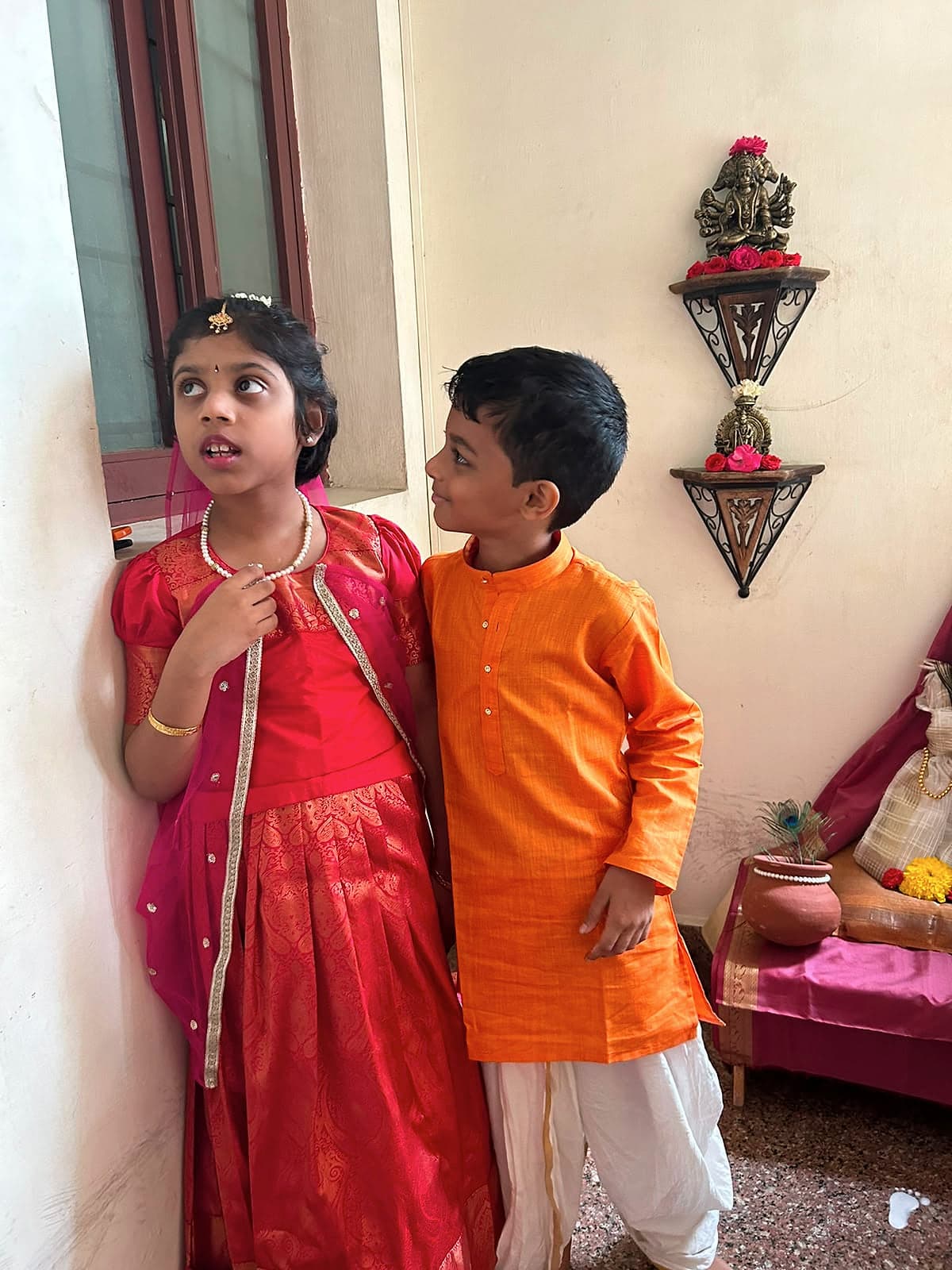 A girl in a red silk skirt and a boy in an orange kurta standing together in festival dress.