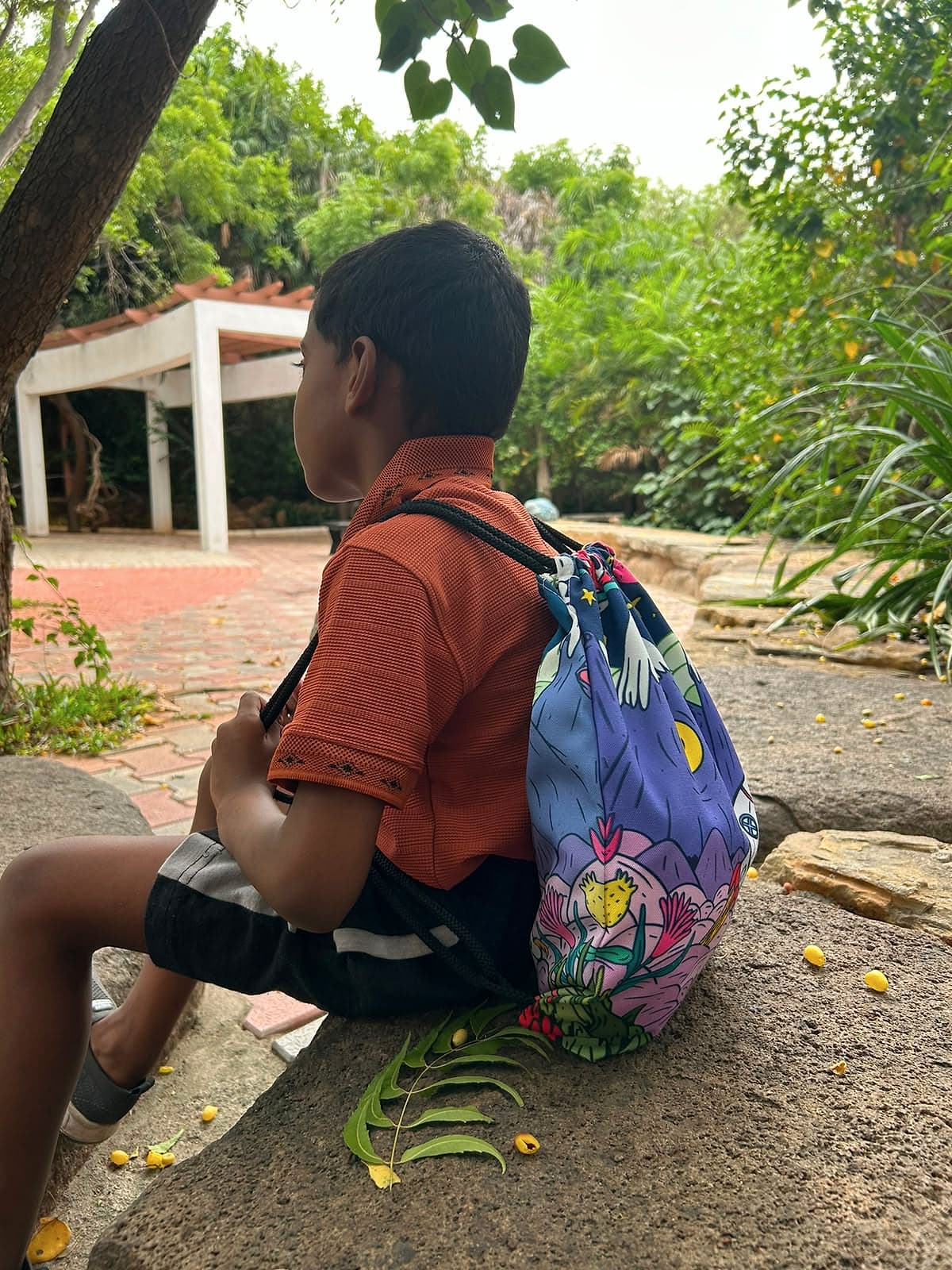 A boy with a patterned drawstring bag sitting on a rock in a leafy garden.