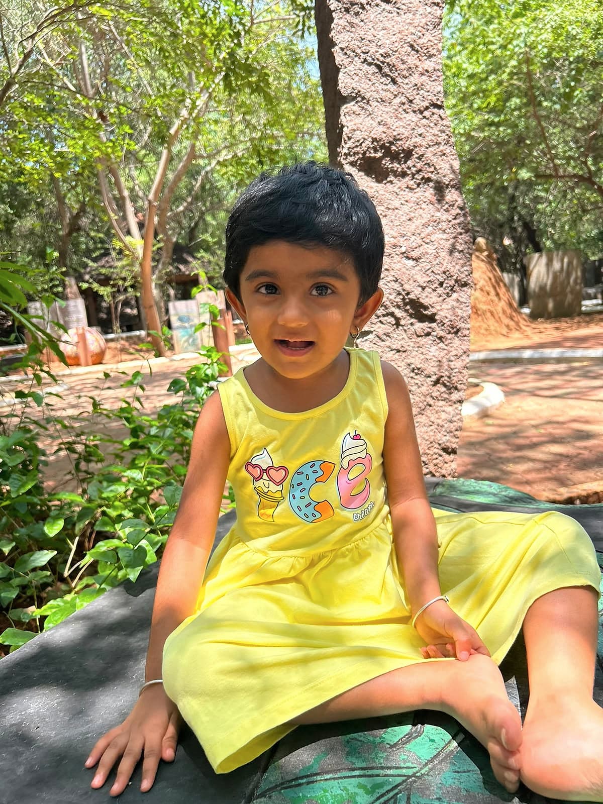 A young child in a yellow dress sitting in the shade of a tree in a park.
