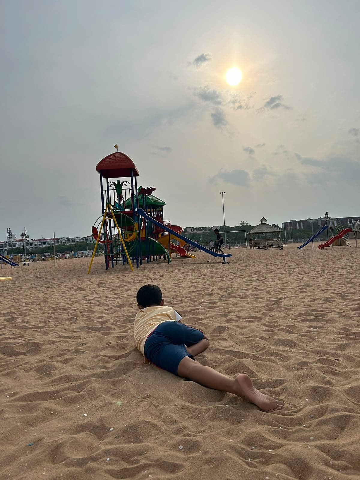A child lying on the sand, looking out toward a playground under a hazy sun.